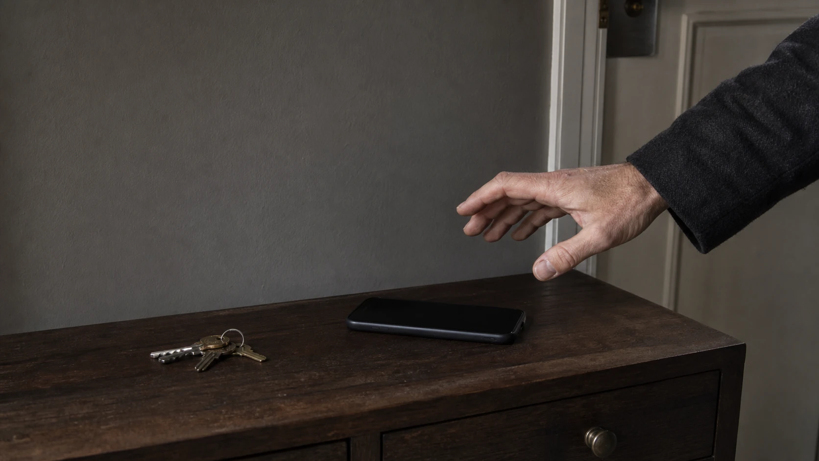 Une main suspendue au-dessus d’un téléphone à l’écran éteint, posé sur une console d’entrée près d’un trousseau de clés.
