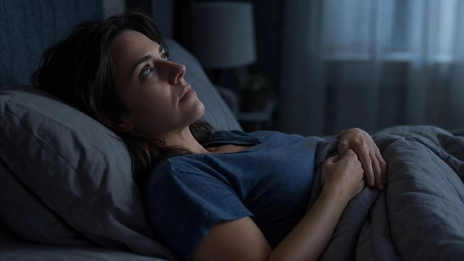 Femme brune allongée dans un lit sombre, regard vers le plafond, mains croisées sur la couverture